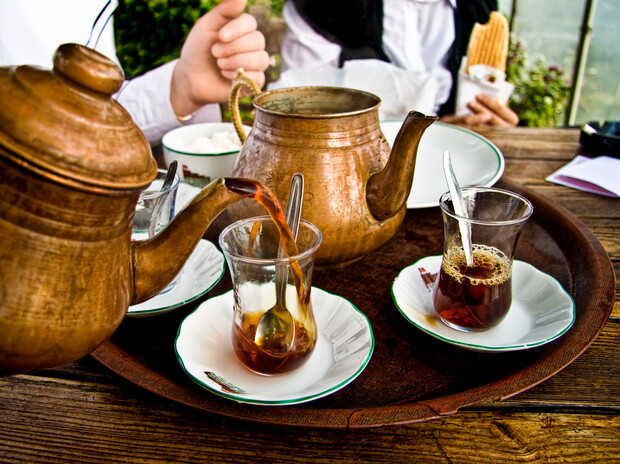 Two copper teapots pour tea into small glasses on a tray, placed on a wooden table. A person in the background holds a cone, suggesting an outdoor café setting.