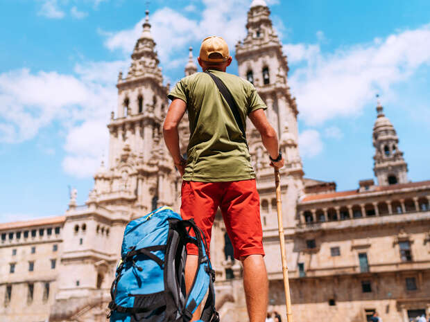 A person with a large blue backpack and walking stick stands facing an ornate, historic cathedral with two tall towers under a clear blue sky.