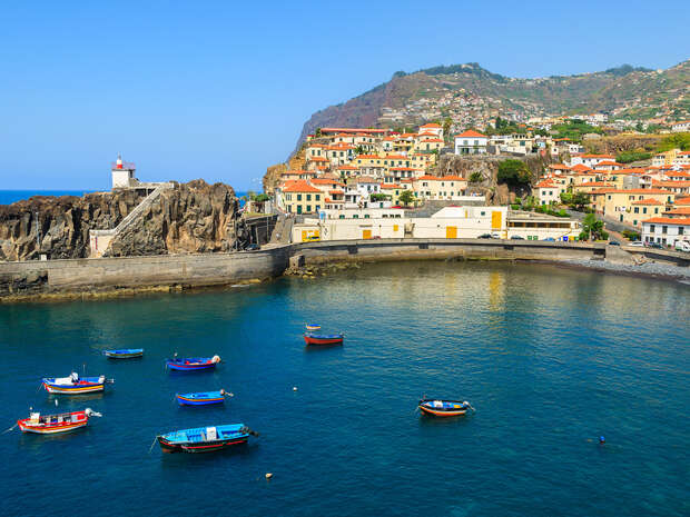 Colorful boats float in a calm, clear blue bay surrounded by a coastal town with vibrant buildings and a rocky lighthouse. Hills with greenery form the backdrop under a clear sky.