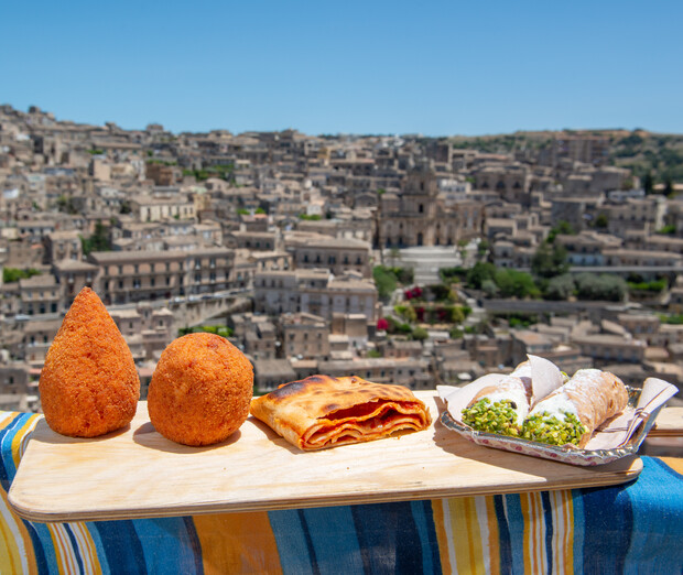 Italian pastries and savory snacks rest on a wooden board atop a colorful cloth, with a sprawling, historic cityscape in the background under a clear blue sky.
