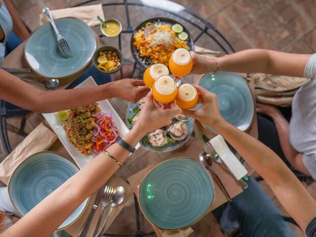 Four hands raise juice glasses in a toast above a table set with plates, various dishes, and silverware, suggesting a shared meal in a social dining setting.