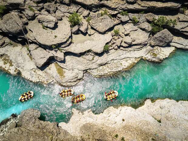 Rafts float along a narrow, turquoise river flanked by rocky cliffs with sparse vegetation. The aerial view captures several groups of people paddling through the winding waterway.