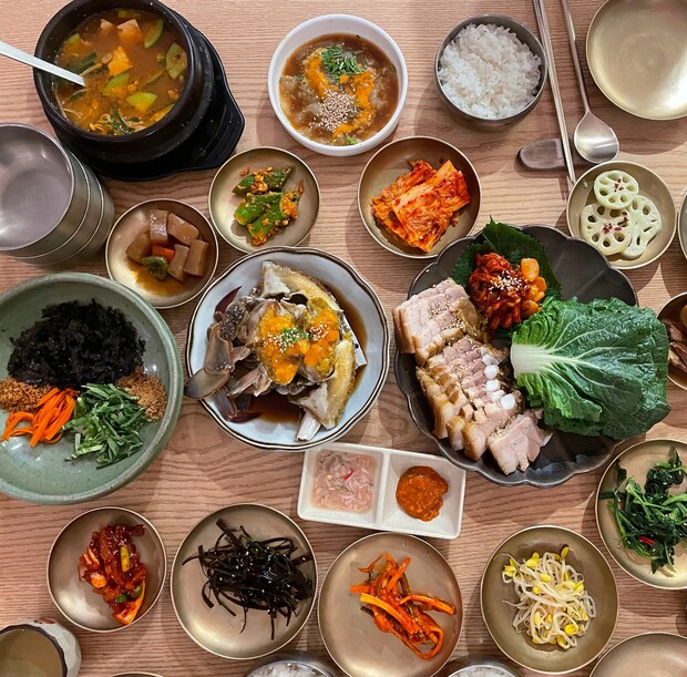 Various dishes are arranged on a wooden table, featuring Korean cuisine including sliced meat, kimchi, soups, and assorted side dishes in individual bowls, accompanied by utensils.