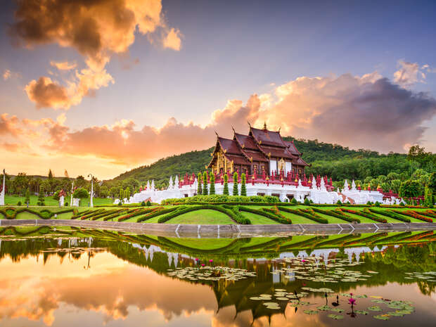 A traditional Thai building stands majestically against a sunset sky, reflecting in a serene pond with lily pads, surrounded by manicured gardens and lush greenery.
