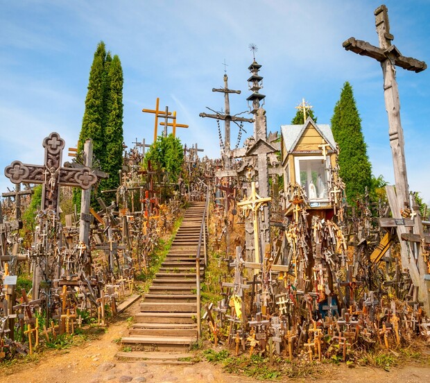 Wooden crosses of various sizes fill a hillside, creating a forest-like scene. A wooden staircase ascends through the middle. Tall, narrow trees flank the area under a blue sky.