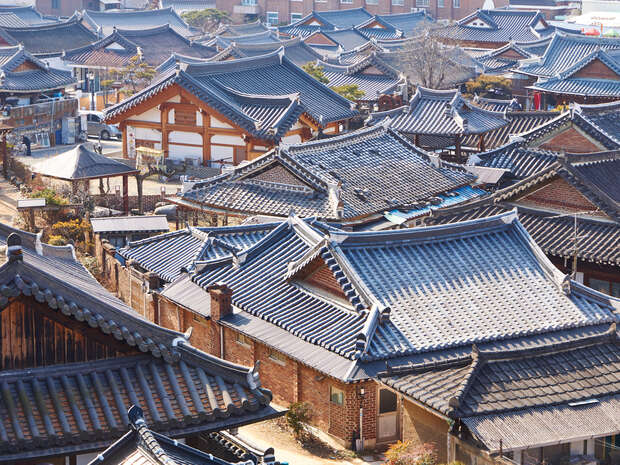 Traditional Korean houses with tiled roofs densely packed together, creating a picturesque village scene. Sunlight highlights the intricate architecture, with bare trees interspersed, set against a backdrop of modern buildings.