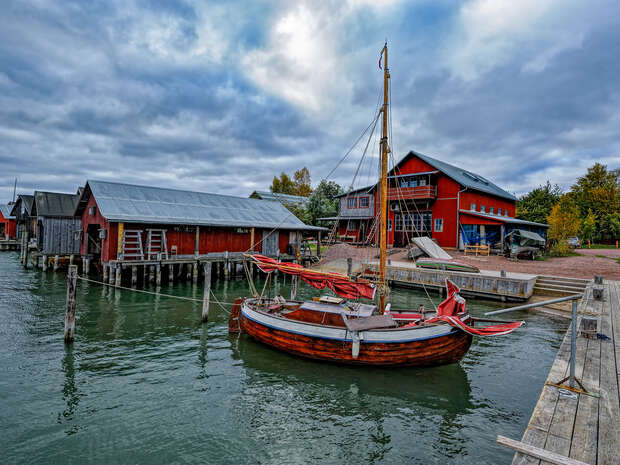 A small wooden sailboat is docked in calm water, surrounded by rustic red boathouses and a cloudy sky, creating a quaint maritime village scene.