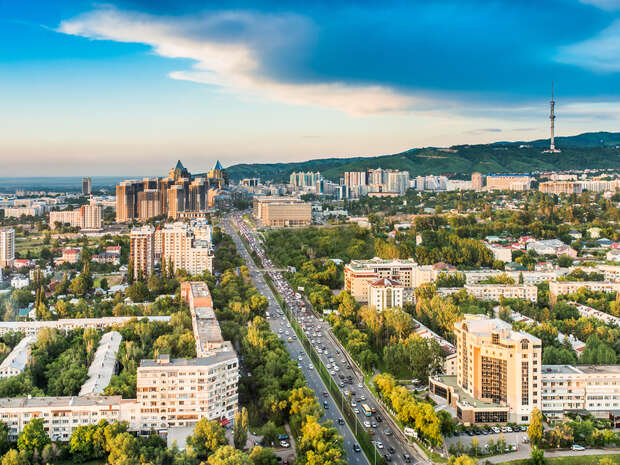 Aerial view of a bustling city with tall buildings and busy roads, surrounded by lush greenery and mountains in the background, under a partly cloudy sky.