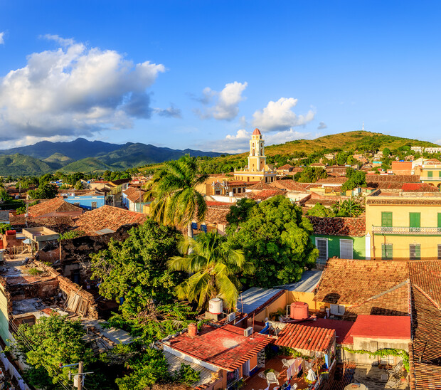 Colorful colonial buildings with red-tiled roofs are nestled among lush green trees, with a prominent church tower rising against a backdrop of distant mountains and a clear blue sky.