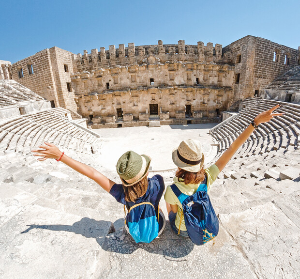 Two people wearing hats sit with arms raised, facing an ancient amphitheater's stone stage. The amphitheater is surrounded by tiered seating and framed by a clear blue sky.