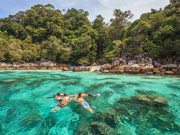 Three people snorkel in clear turquoise water near the shore, surrounded by rocks and a dense, green forest with a small boat docked on the sandy beach.