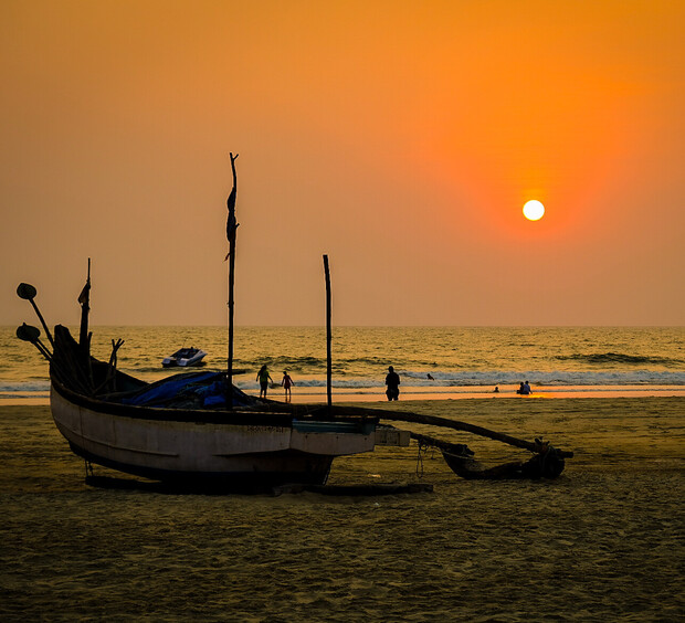 A weathered boat rests on a sandy beach as the sun sets over calm ocean waves, silhouetting people walking and swimming in the warm orange glow of the evening sky.