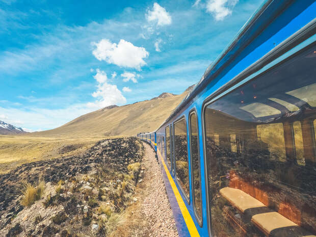A blue train travels on a rocky track through a mountainous landscape, reflecting the sky and terrain in its windows. Fluffy clouds dot the clear, blue sky above.