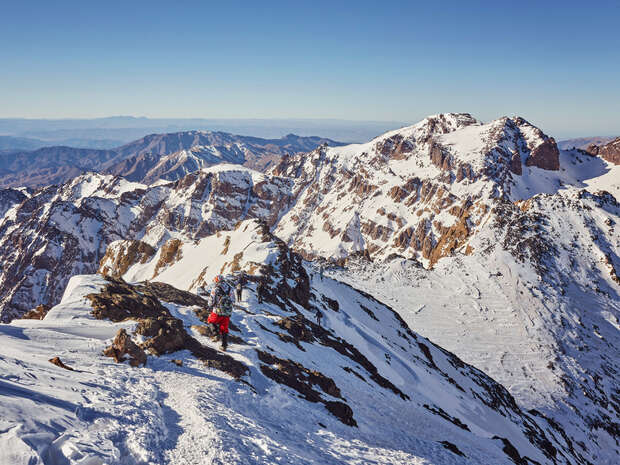 Climbers ascend a snow-covered mountain ridge with rocky peaks towering around them, under a clear blue sky, creating a dramatic and adventurous alpine vista.