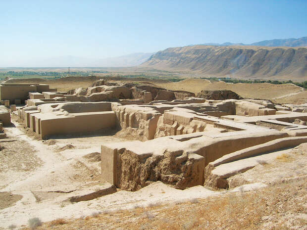 Ancient ruins form rectangular, sun-baked mud structures amidst a desert landscape. In the background, distant mountains rise under a clear blue sky, overlooking a sparse, arid terrain.