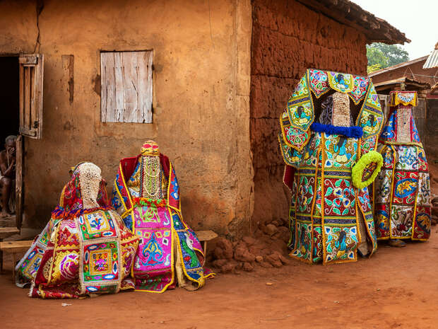 Colorful, elaborately dressed figures adorned in intricate beaded and embroidered costumes stand and sit outside a rustic, clay-walled building, exuding a traditional, cultural ceremonial ambiance.