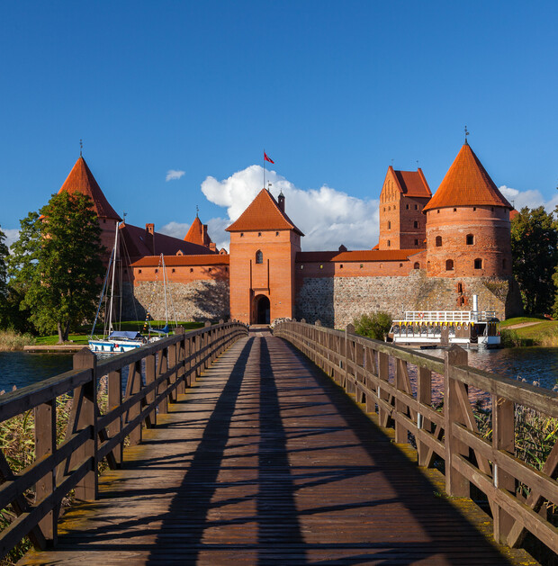 A red-brick castle with conical towers stands under a blue sky. A wooden bridge leads to the entrance, surrounded by water and moored boats. Trees frame the scene.