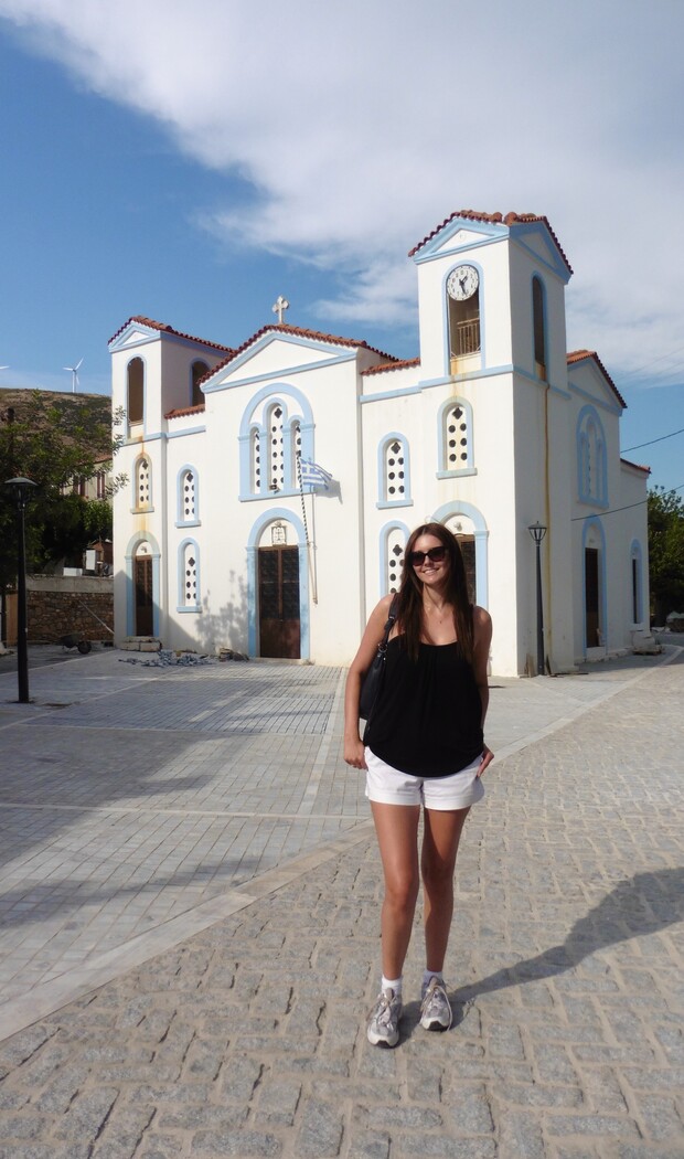 A person stands smiling in front of a large, white church with blue trim and a clock tower. A Greek flag is visible, and the paved area is sunlit.