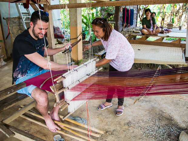A man weaves fabric on a loom with guidance from a woman, seated in a rustic setting. Two people observe in the background, surrounded by wooden structures and greenery.