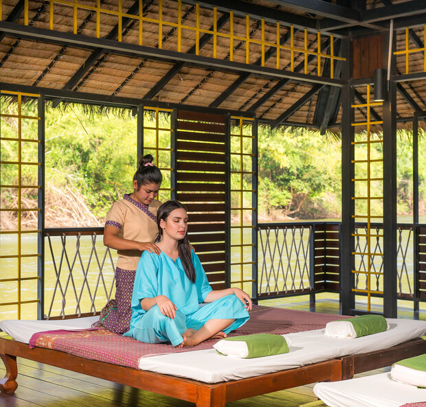 A woman sits cross-legged receiving a shoulder massage from another woman in a tranquil, open-air setting with wooden decor and a view of greenery outside.