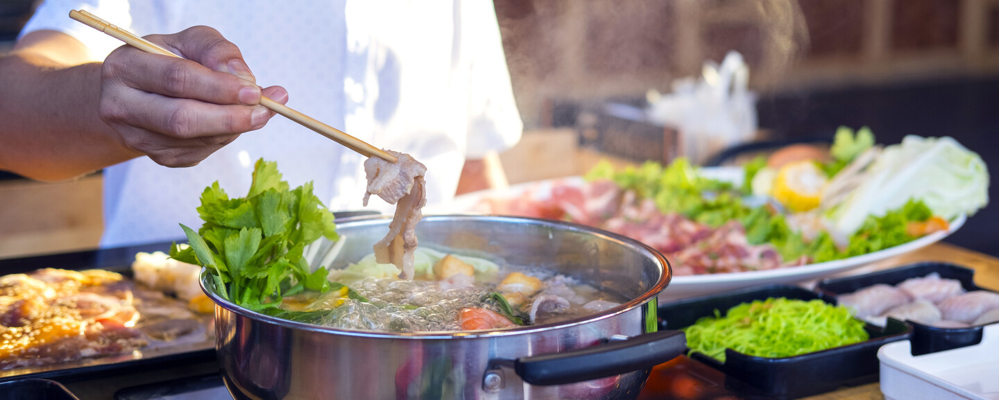 A person uses chopsticks to cook meat in a steaming hot pot with vegetables, surrounded by plates of raw ingredients on a wooden table.