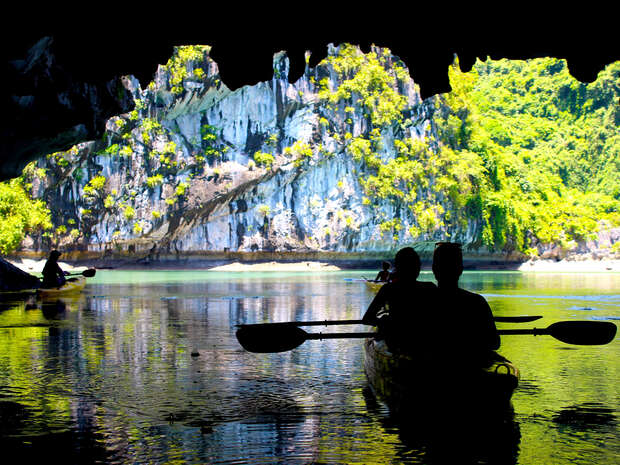 Paddlers navigate a kayak through a cave opening, entering a sunlit lagoon surrounded by lush, green cliffs, under a bright blue sky.