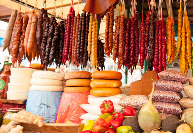 Various colorful sausages hang on strings above round cheeses and fresh vegetables at a market stall filled with vibrant produce and goods.