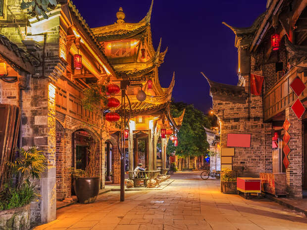 Traditional Chinese buildings adorned with red lanterns stand illuminated at night, lining a cobblestone street in a quiet, historic district. A bicycle leans against a wall.