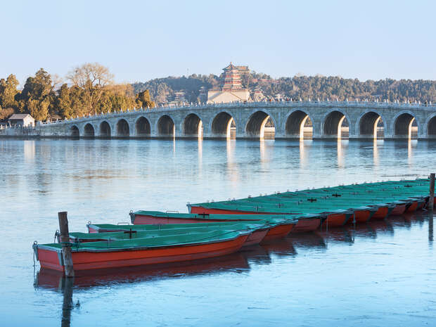 Red and green boats are moored on a calm lake in front of an arched stone bridge. Behind the bridge, a historical building sits amidst a forested hillside under a clear sky.