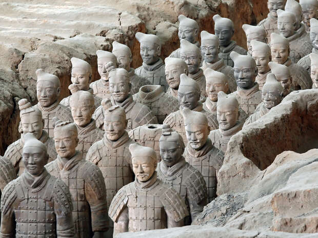 Terracotta soldiers stand in rows, displaying unique armor and headgear. They are positioned in an excavation pit, surrounded by earthen walls, representing Qin Shi Huang's mausoleum army.
