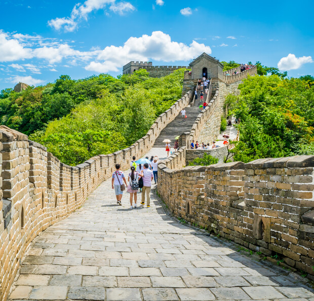 Stone wall stretches upward with people walking along it; lush green trees surround the structure under a bright blue sky with fluffy clouds.