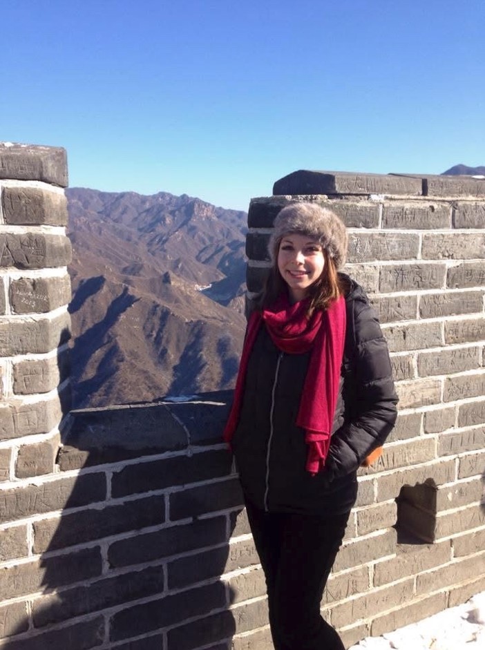 A person wearing a winter coat and hat stands against a stone wall, with distant brown mountain ranges under a clear blue sky.