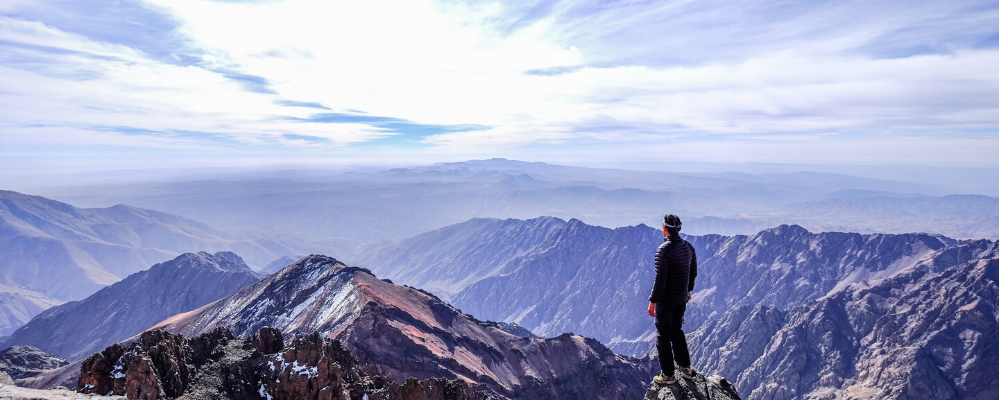 A person stands on a rocky mountain peak, gazing at expansive, layered mountain ranges under a bright sky with scattered clouds.