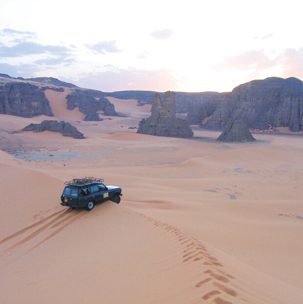 A rugged vehicle ascends a sandy dune, leaving tracks, surrounded by vast desert and rocky formations under a pastel sunset sky.