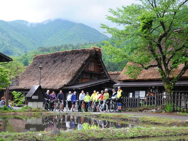 Cyclists stand with their bikes beside a reflective pond in front of traditional thatched-roof houses, surrounded by green trees and mountains under a cloudy sky.