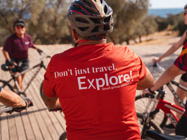 Cyclist wearing a red shirt with text "Don't just travel. Explore! Small group adventures." gathers with others on a sunny, wooden path surrounded by trees and distant water.