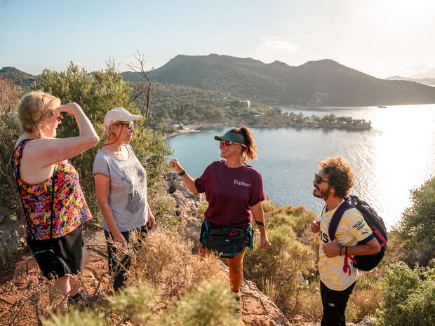 Four people stand on a rocky, shrub-covered hill overlooking a scenic landscape with a body of water and mountains. One person gestures while interacting with others. Bright daylight illuminates the scene.