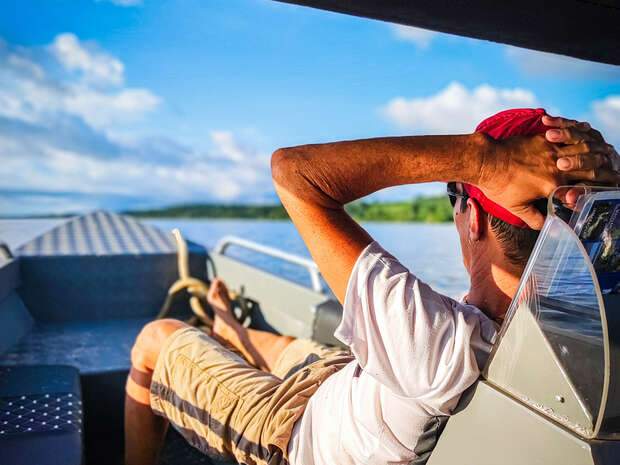 A person relaxes on a boat, resting with hands behind their head and feet up. They wear a red cap and sunglasses, surrounded by calm water and a distant shoreline.