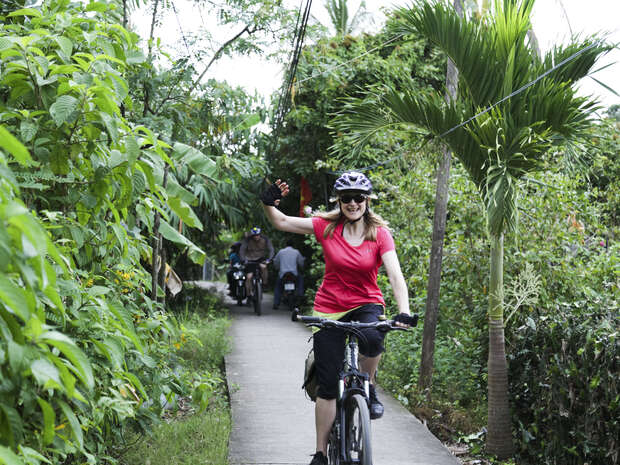 A person rides a bicycle on a narrow path, surrounded by lush greenery and palm trees. Behind, another cyclist follows. The person in front waves and smiles, wearing a helmet.