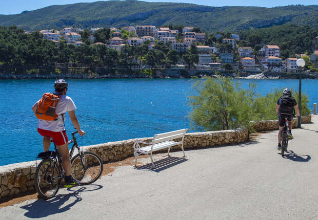 Cyclists ride along a seaside path beside a stone wall, overlooking blue water. Houses and green hills are visible in the background. A white bench stands nearby under a clear sky.
