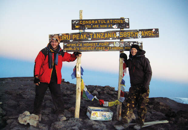 Two people stand beside a wooden sign on rocky terrain. The sign reads: “CONGRATULATIONS YOU ARE NOW AT UHURU PEAK, TANZANIA, 5895 M AMSL, AFRICA’S HIGHEST POINT, WORLD'S HIGHEST FREE-STANDING MOUNTAIN.”