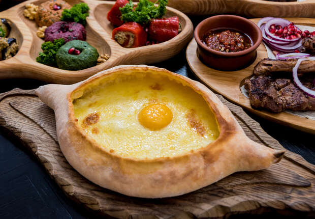 A boat-shaped bread topped with melted cheese and a sunny-side-up egg sits on a wooden board, surrounded by colorful dips, roasted vegetables, and grilled meat garnished with onions.