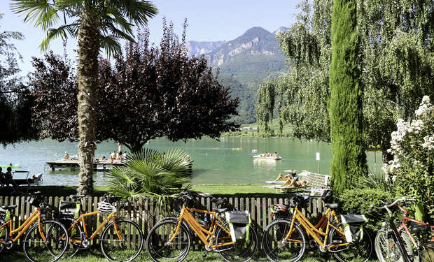 Bright orange bicycles are parked in front of a scenic lake with people relaxing on docks and pedaling boats, surrounded by lush green trees and mountains in the background.