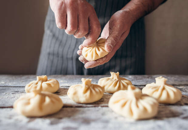 Hands with flour dust sculpt dumplings on a wooden table, surrounded by several neatly arranged, uncooked dough parcels.