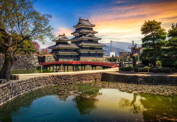 A traditional Japanese castle stands surrounded by a stone wall and calm water, with a red arched bridge leading to it. Trees and mountains are visible under a vibrant sunset sky.