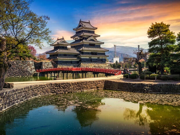 A traditional Japanese castle stands beside a red arched bridge, reflected in a calm pond. Surrounded by trees and stone walls, mountains are visible under a colorful sunset sky.