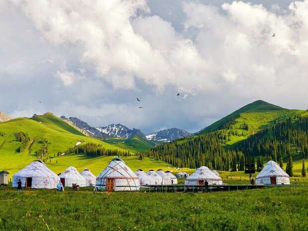 Yurts stand in a green valley surrounded by rolling hills and distant mountains, under a cloudy sky with birds flying above, creating a serene, natural setting.