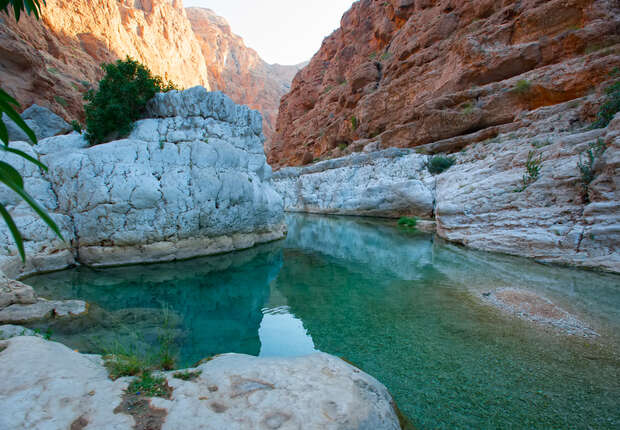 Clear turquoise water flows gently between rocky, canyon walls. White and reddish-brown rocks surround the calm, inviting pool. Vegetation grows sparsely on the rugged cliffs under a bright blue sky.