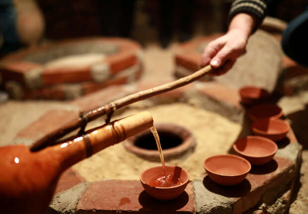 A clay pitcher pours liquid into small clay bowls using a stick with a spout, surrounded by a rustic brick and sand setup, suggesting a traditional setting.