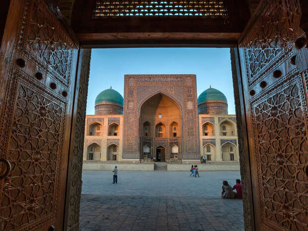 An intricately decorated wooden door opens to reveal a large courtyard with a grand, arched building featuring turquoise domes, while people walk and sit in the plaza.